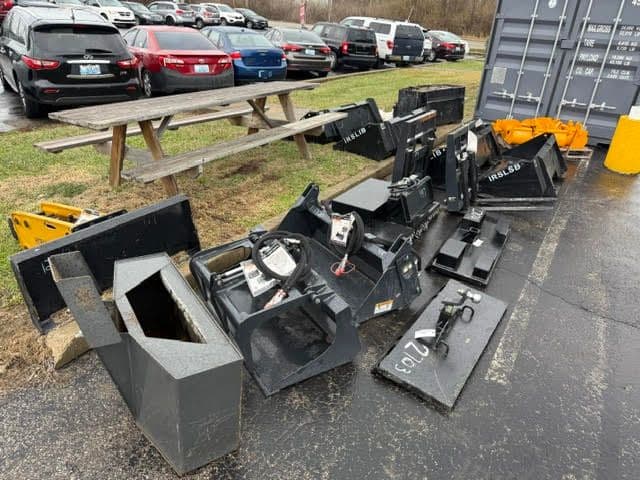 Various heavy equipment attachments arranged on a pavement near a picnic table and parked vehicles.