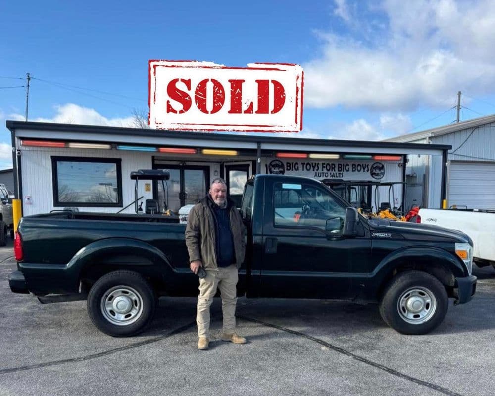 Man standing next to a sold green pickup truck outside an auto sales shop.