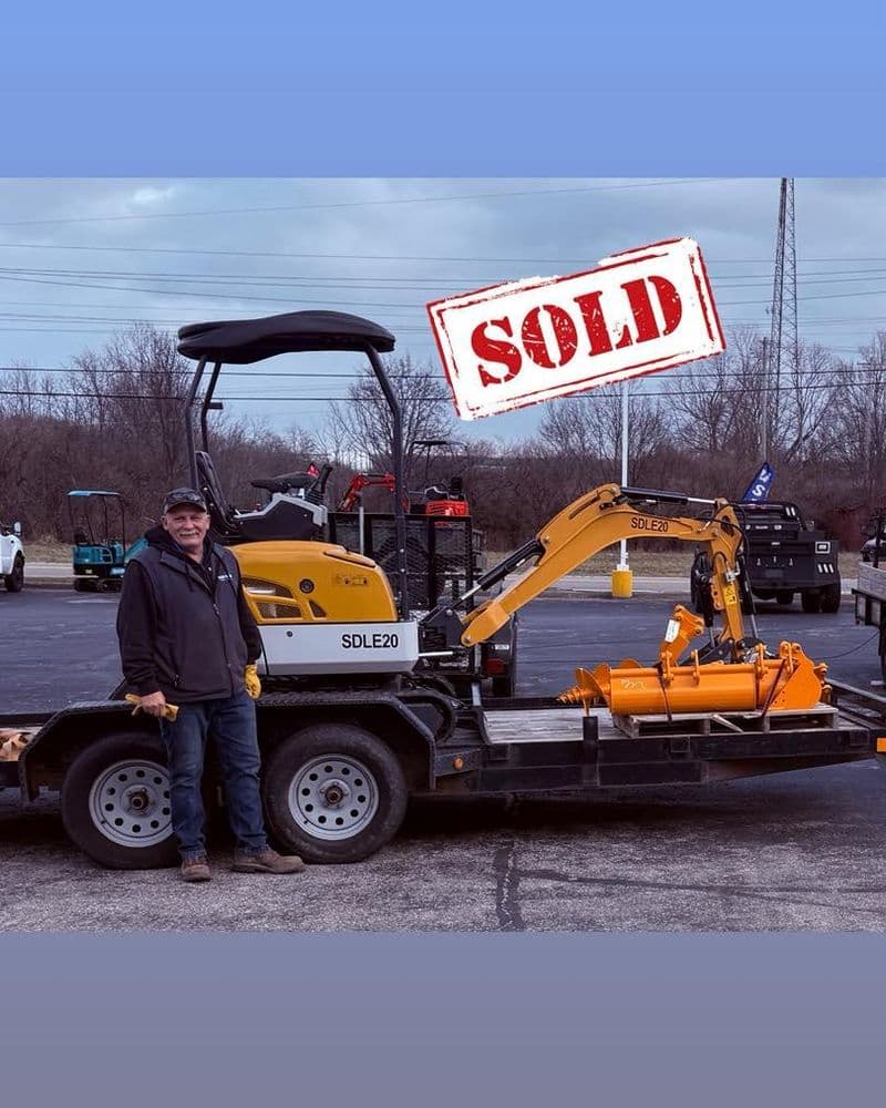 Man standing next to a sold SDLE20 mini excavator on a trailer with an industrial background.