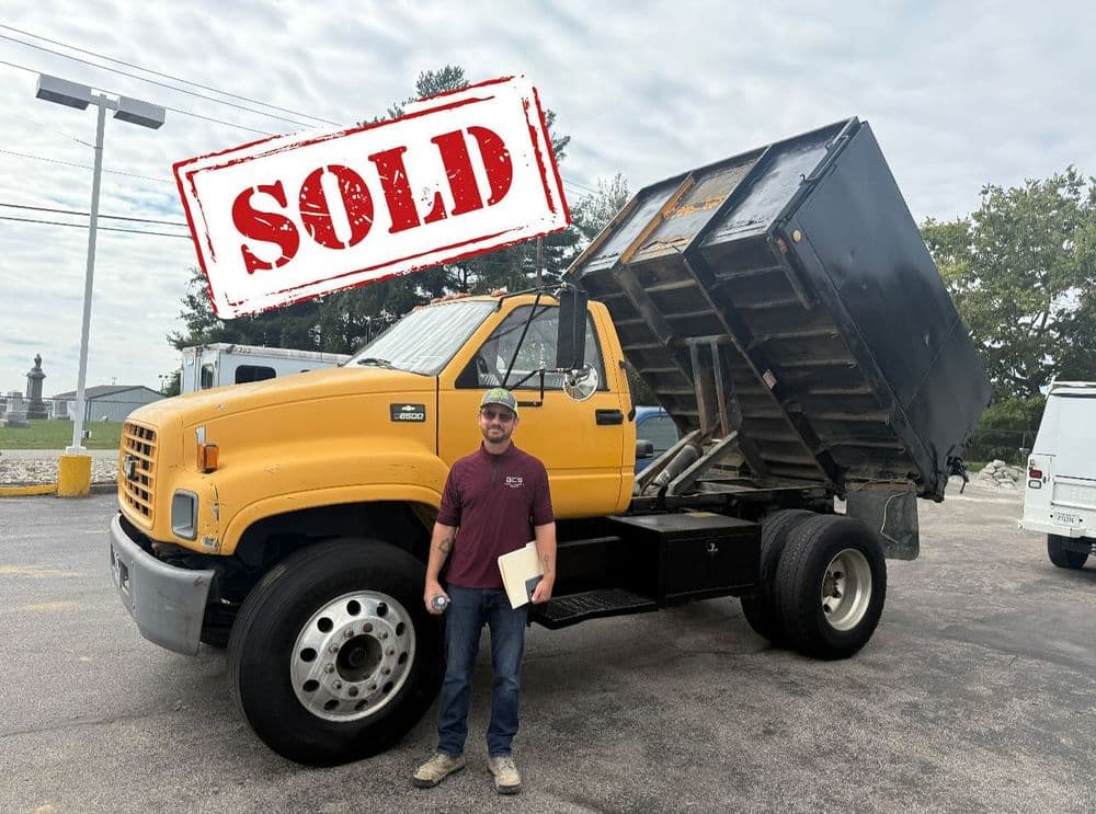 Yellow dump truck with sold stamp and man holding documents outdoors.