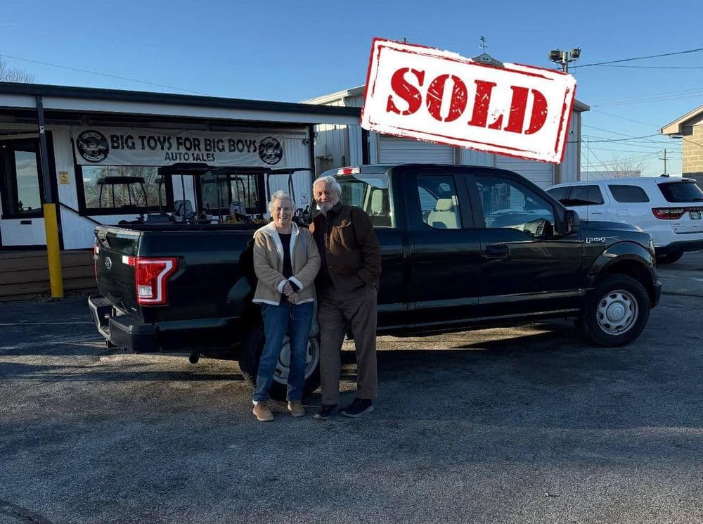 Happy couple standing beside a sold black pickup truck outside an auto sales dealership.
