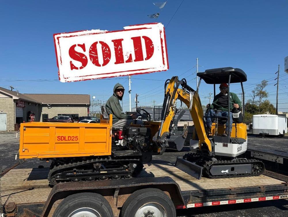 Mini excavator on trailer marked 'SOLD', with two workers in hoodies against a blue sky.