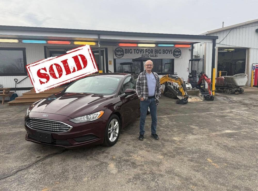 Man standing next to sold maroon Ford Fusion in front of Big Toys for Big Boys auto sales.
