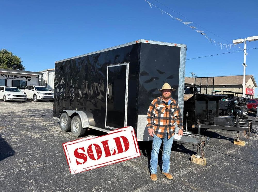 Man in plaid shirt stands next to a sold black trailer under a clear blue sky.
