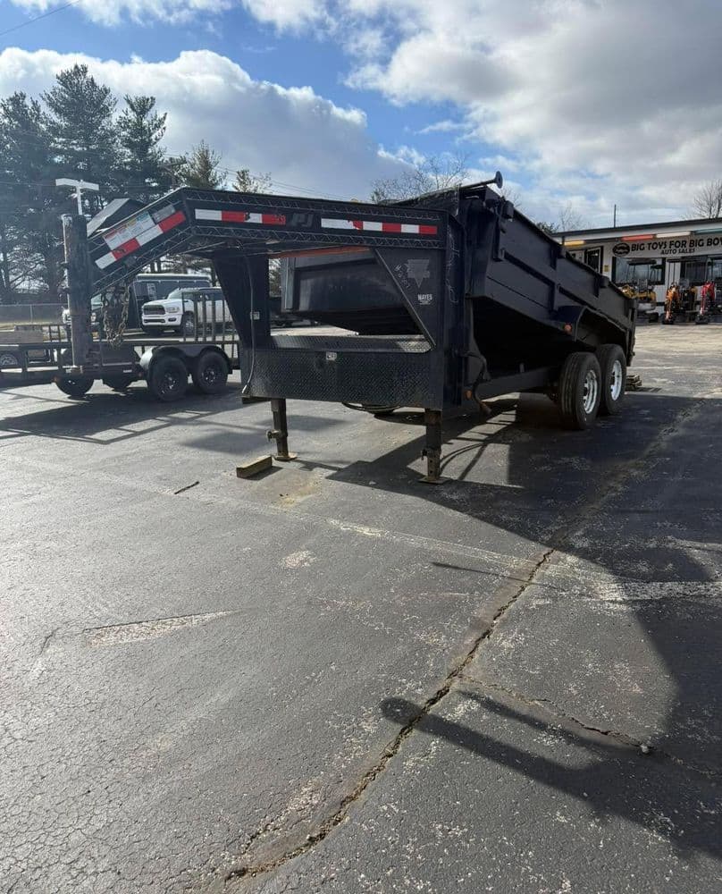 Black dump trailer parked in a lot, with a clear blue sky and trees in the background.