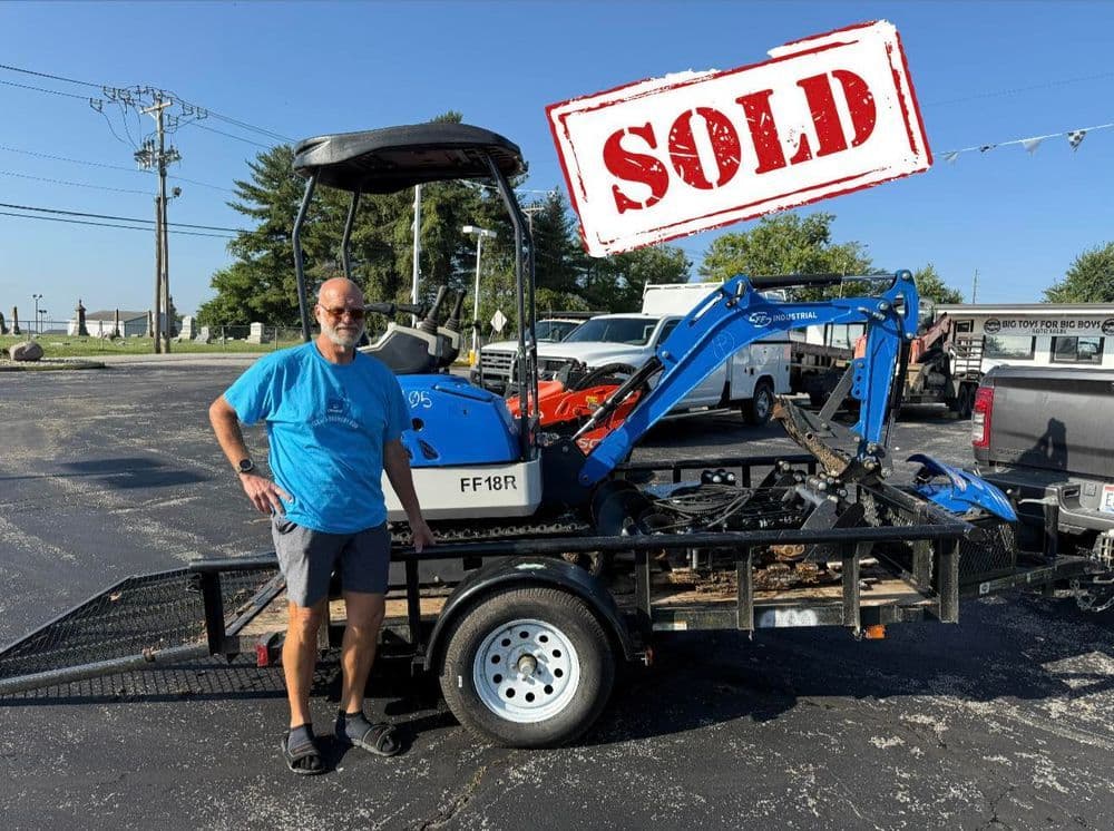 Man standing next to a sold mini excavator on a trailer in a parking lot.