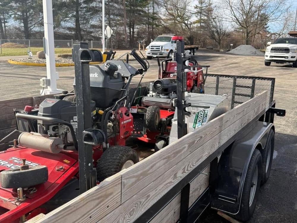 Red lawn mowers loaded on a trailer in a outdoor setting with parked trucks nearby.