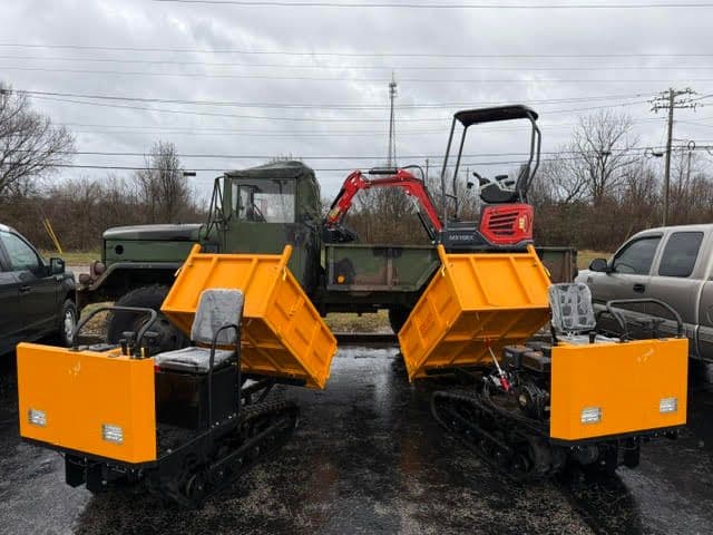 Two compact tracked snow plows with yellow blades, parked next to vehicles in a cloudy setting.