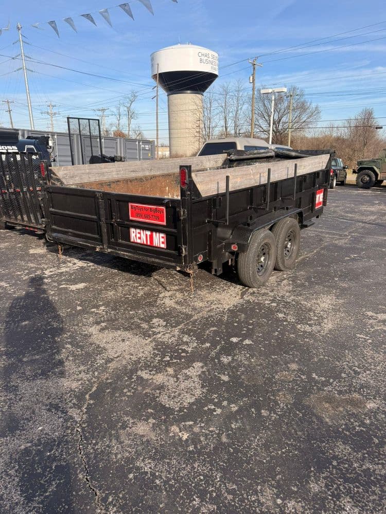 Black trailer for rent with "RENT ME" sign, parked on a gravel lot alongside equipment.