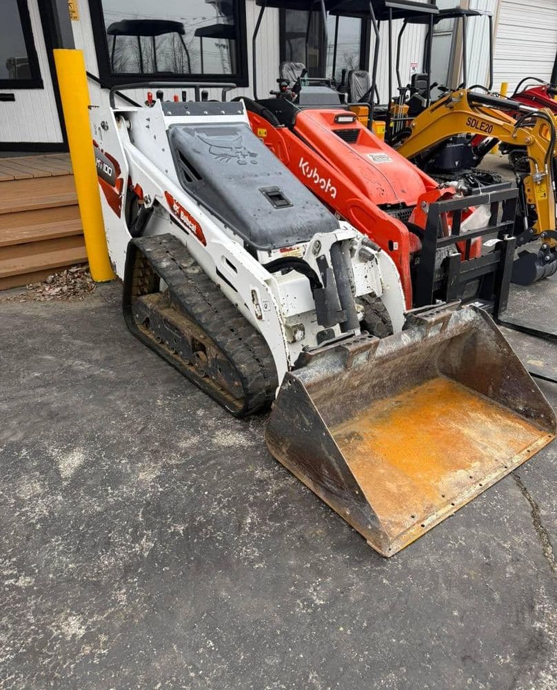 Compact track loader with a bucket attachment, parked near heavy machinery for construction use.