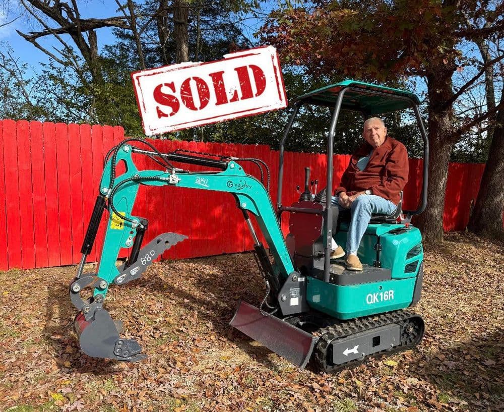 Man sitting on a sold mini excavator in a wooded area with a red fence background.