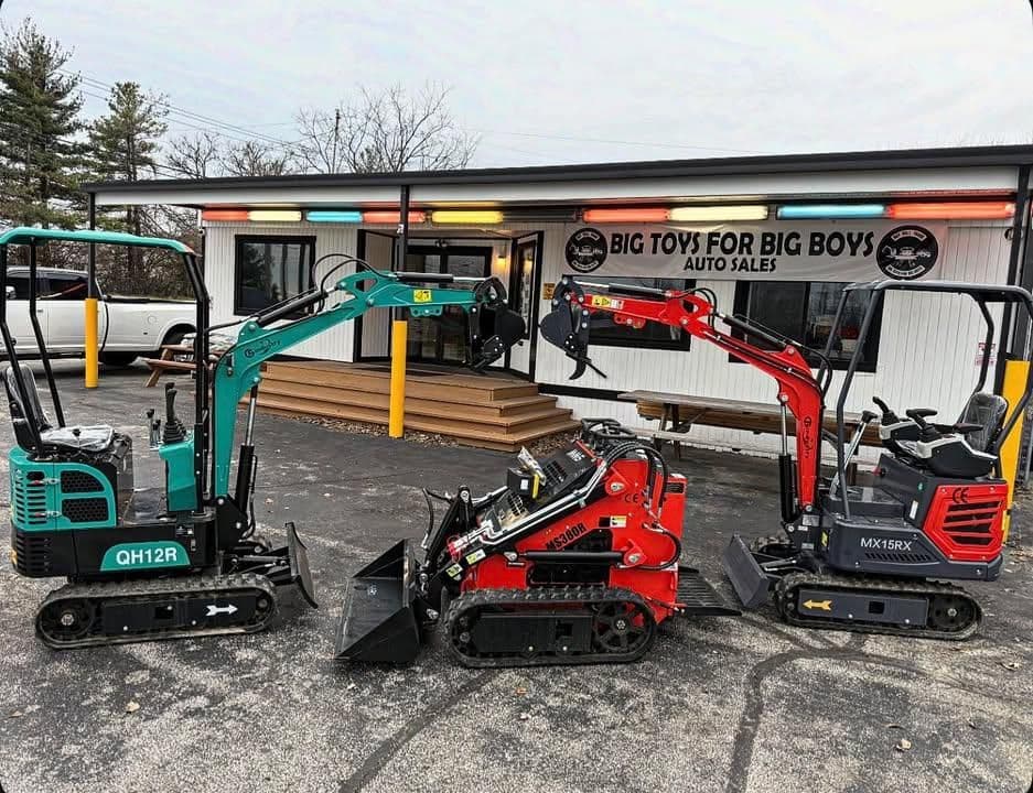 Compact excavators displayed outside "Big Toys for Big Boys" auto sales, showcasing vibrant colors.