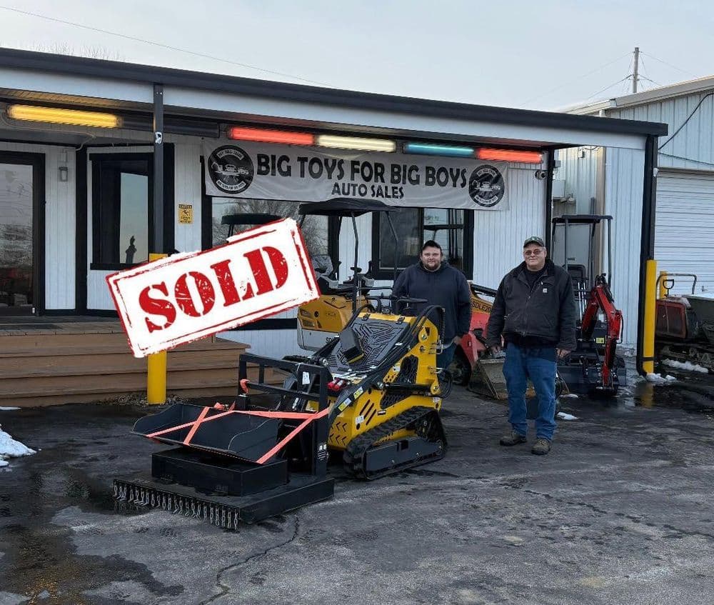 Two men stand in front of Big Toys for Big Boys Auto Sales with a sold compact track loader.