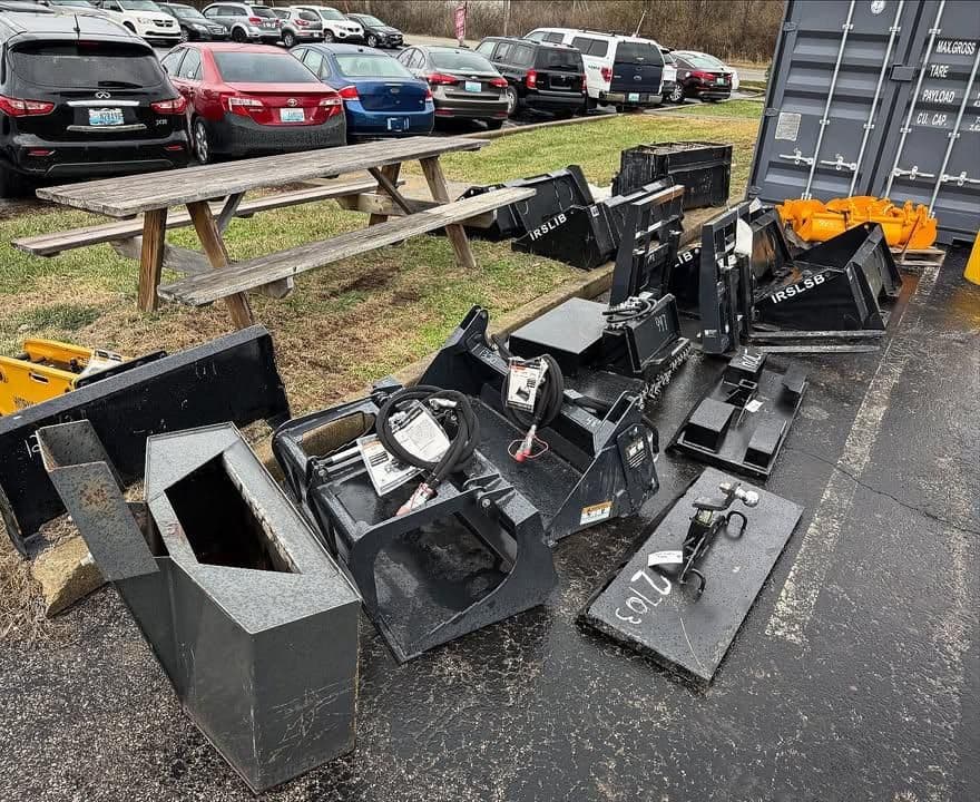 Various heavy equipment attachments arranged outdoors near parked vehicles and a shipping container.